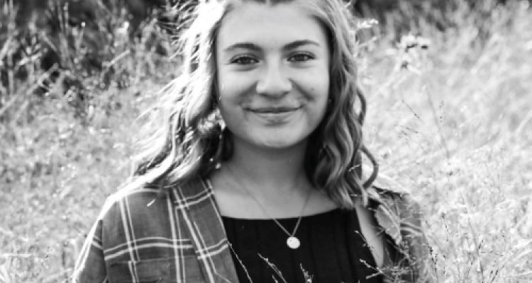 Emily Yanchak headshot. Black and white photo of girl standing in a field of tall grass.